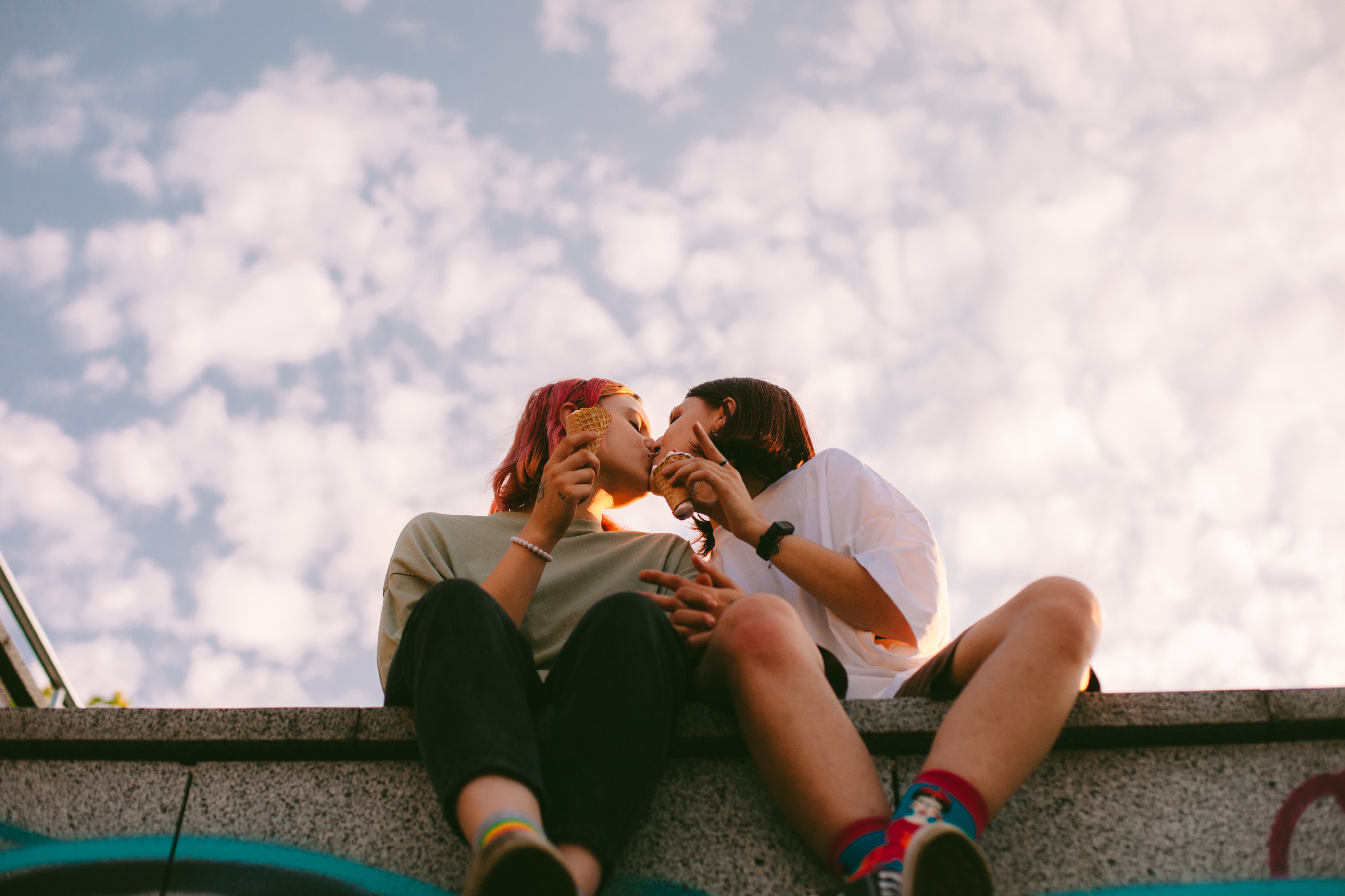 Two people sitting on a ledge outdoors, sharing a kiss while holding ice cream cones, under a partly cloudy sky