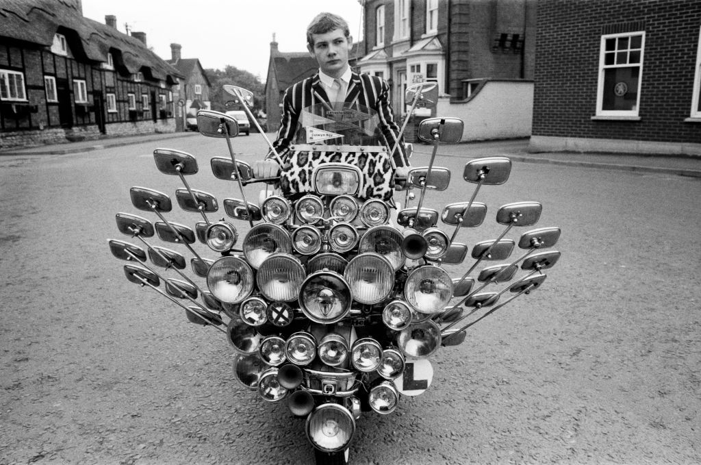 Person riding a scooter with many mirrors and lights on the front, wearing a striped jacket, in an urban setting