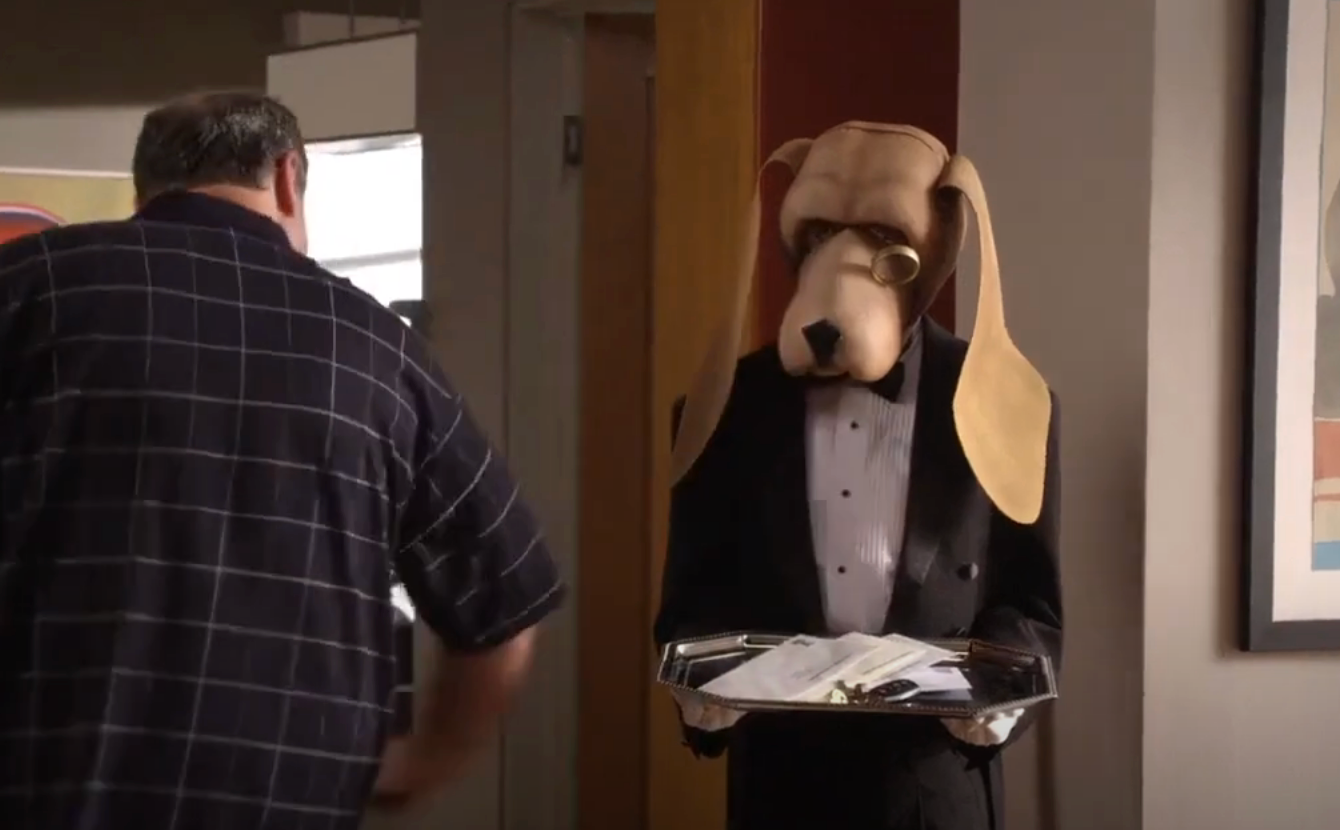 Person in a dog costume wearing a tuxedo holds a tray with papers, interacting with someone in a casual outfit indoors