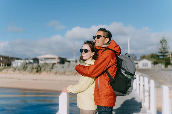 Couple in outdoor jackets and sunglasses smiling, standing on a boardwalk, with arms around each other, looking out to a seaside view