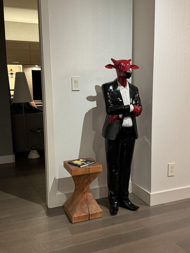 Mannequin with a red bull head in a tuxedo stands beside a small table with a book in a modern room