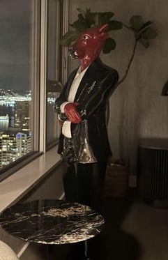A window overlooks a cityscape at night. A devil statue in a suit stands indoors beside a table, adding a quirky element to the modern setting