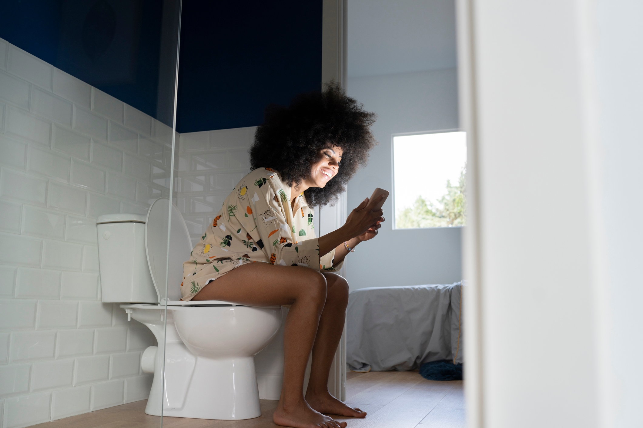 Personen som sitter på et toalett på et moderne bad, smiler på telefonen sin og har på seg en uformell mønstret skjorte. Naturlig lys kommer inn gjennom et vindu