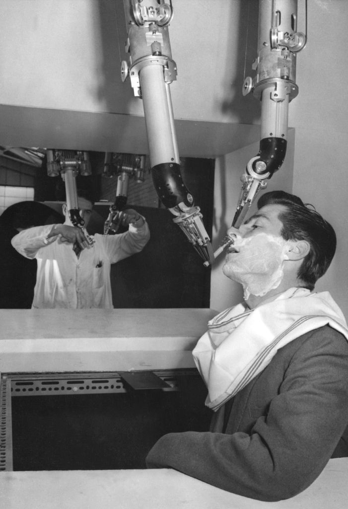 A man receives a shave from robotic arms controlled by a person in a lab coat, illustrating a futuristic grooming concept