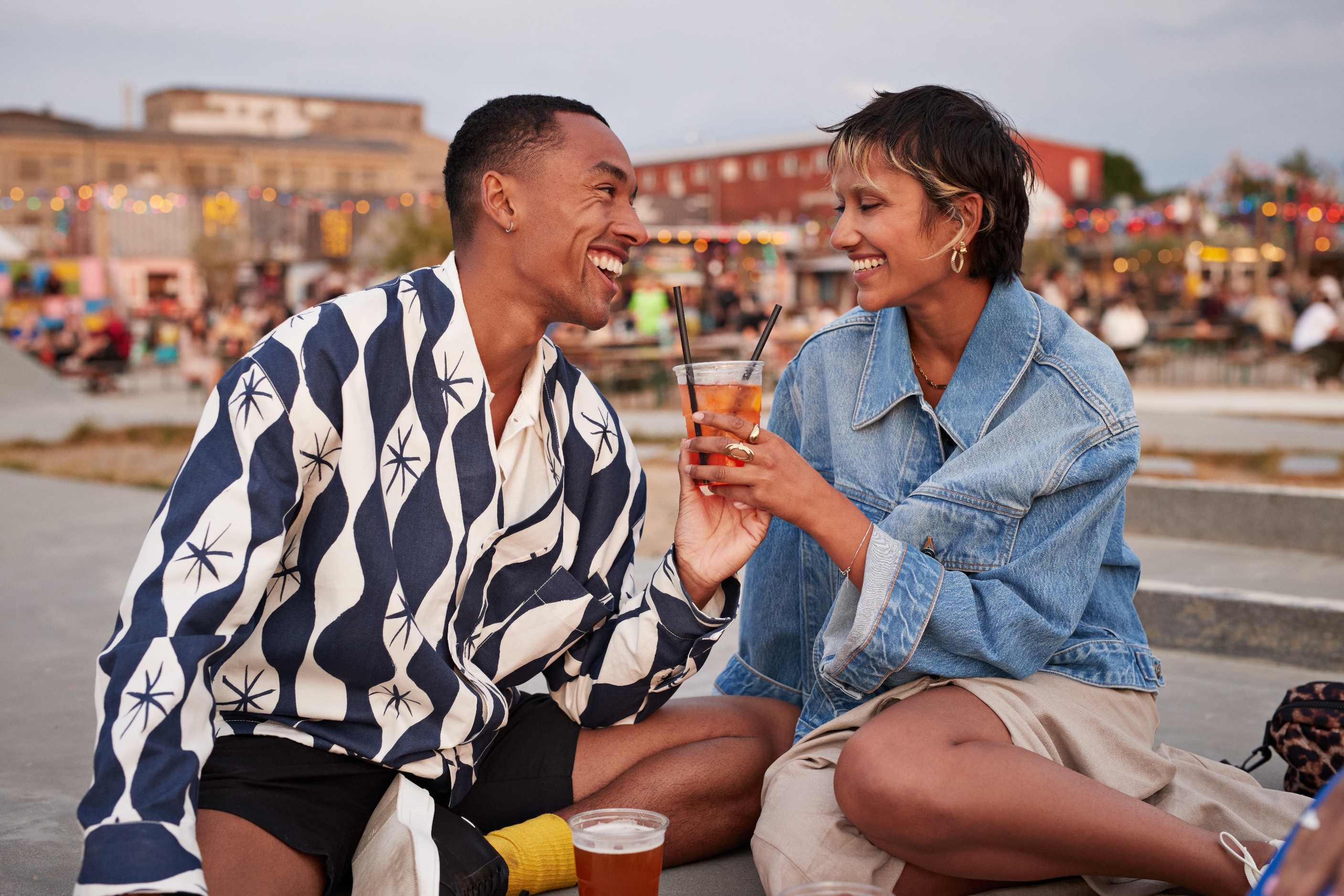 A couple sitting outdoors, smiling and clinking glasses, dressed in casual patterned and denim shirts, enjoying a relaxed and happy moment together