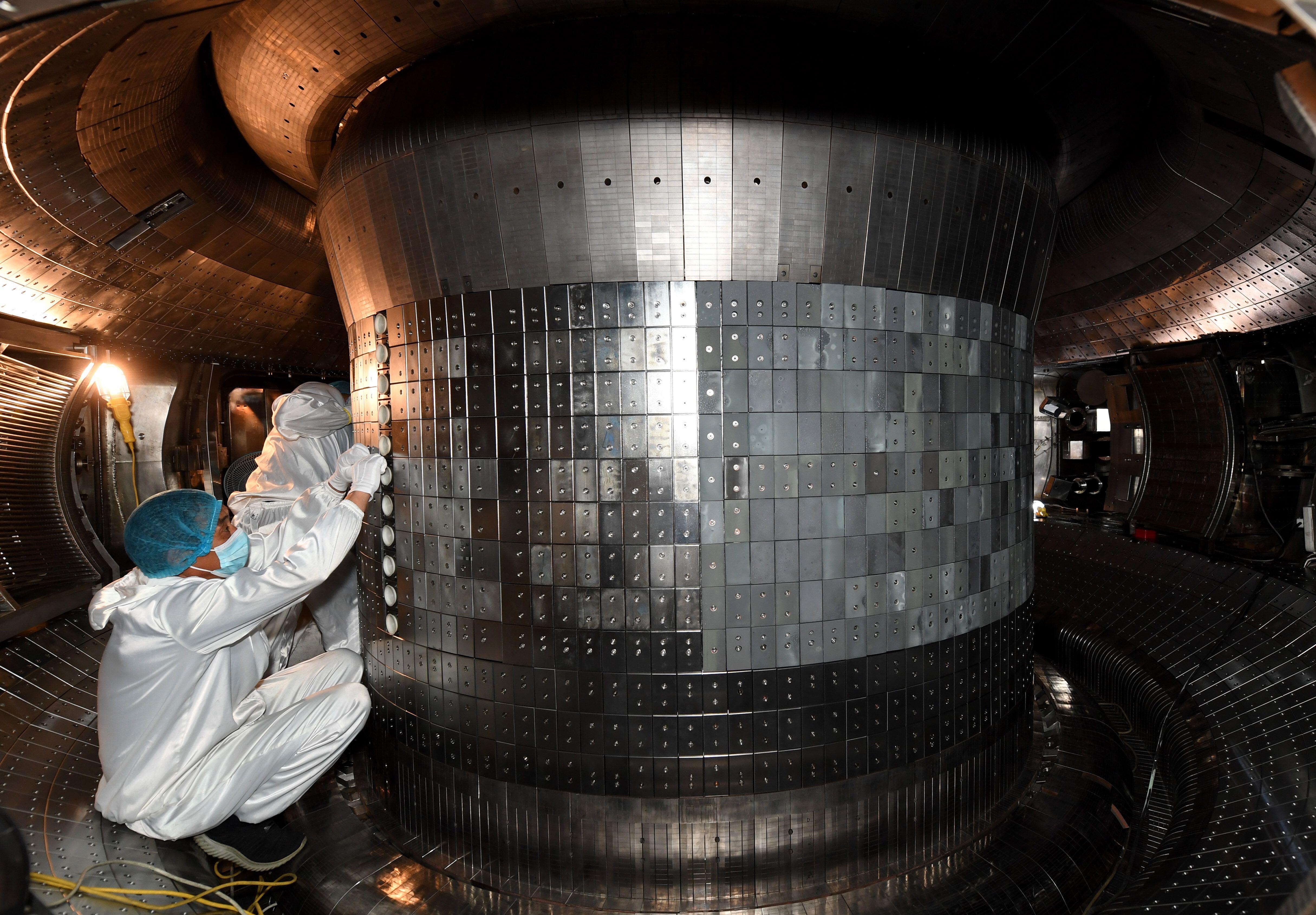 Two people in cleanroom suits inspect a large metallic chamber, indicative of a high-tech or scientific environment