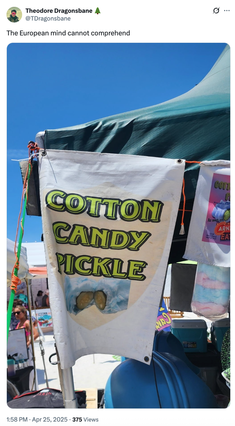 Sign reading "Cotton Candy Pickle" with a pickle in cotton candy on it, under a tent at an outdoor event