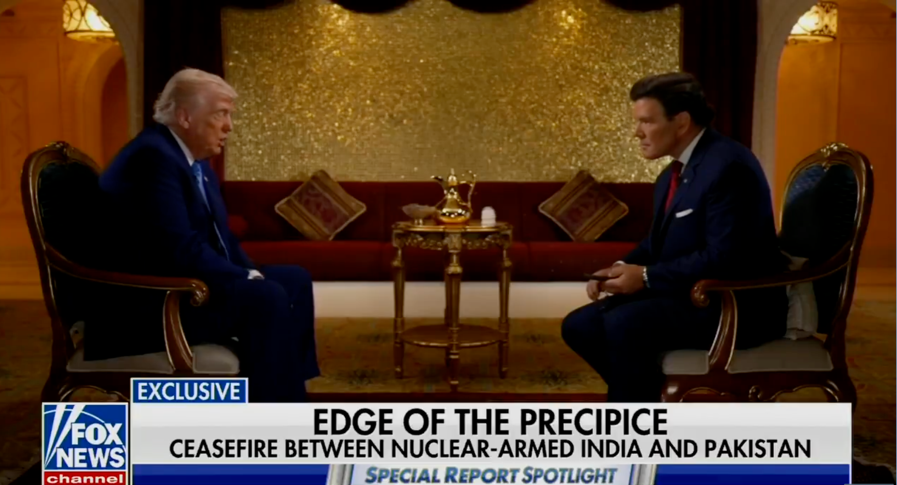 Two men in suits sit facing each other in a formal interview setting. News headlines discuss India-Pakistan ceasefire