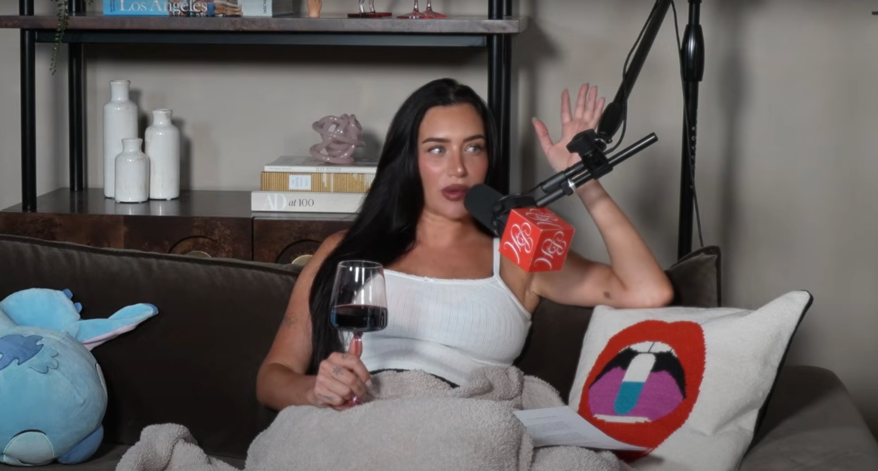 Person with a microphone, sitting on a couch, holding a wine glass, and wrapped in a blanket. Shelves with books and decor in the background