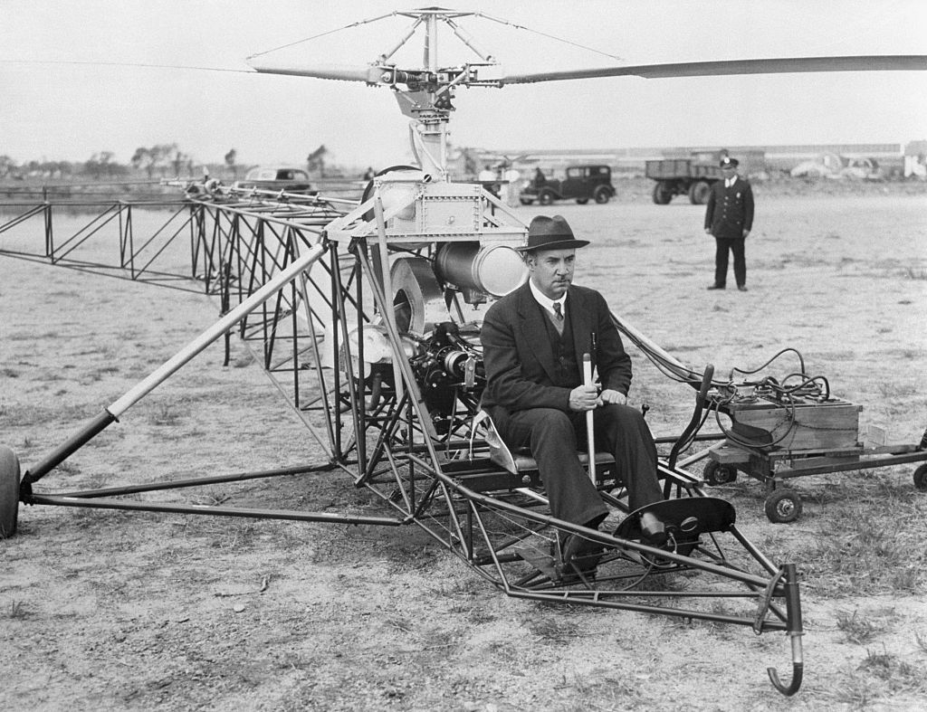 A man in a suit and hat sits on an early helicopter prototype, with visible exposed metal framework, in a field. Another person stands nearby