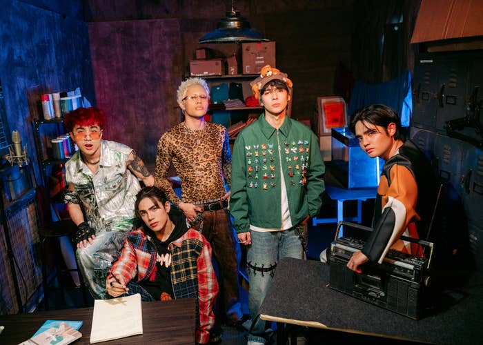 Five individuals in eclectic, fashion-forward outfits pose indoors. One wears leopard print and another a studded green jacket. A boombox is visible