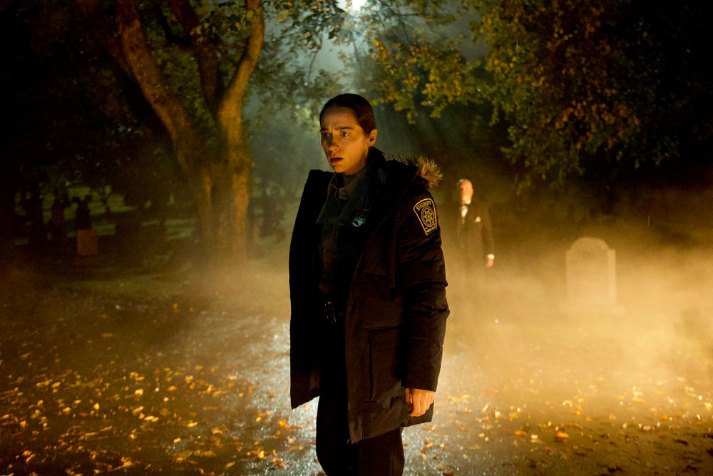 A person in a police uniform stands in a misty, dimly lit graveyard at night, with another figure in the background