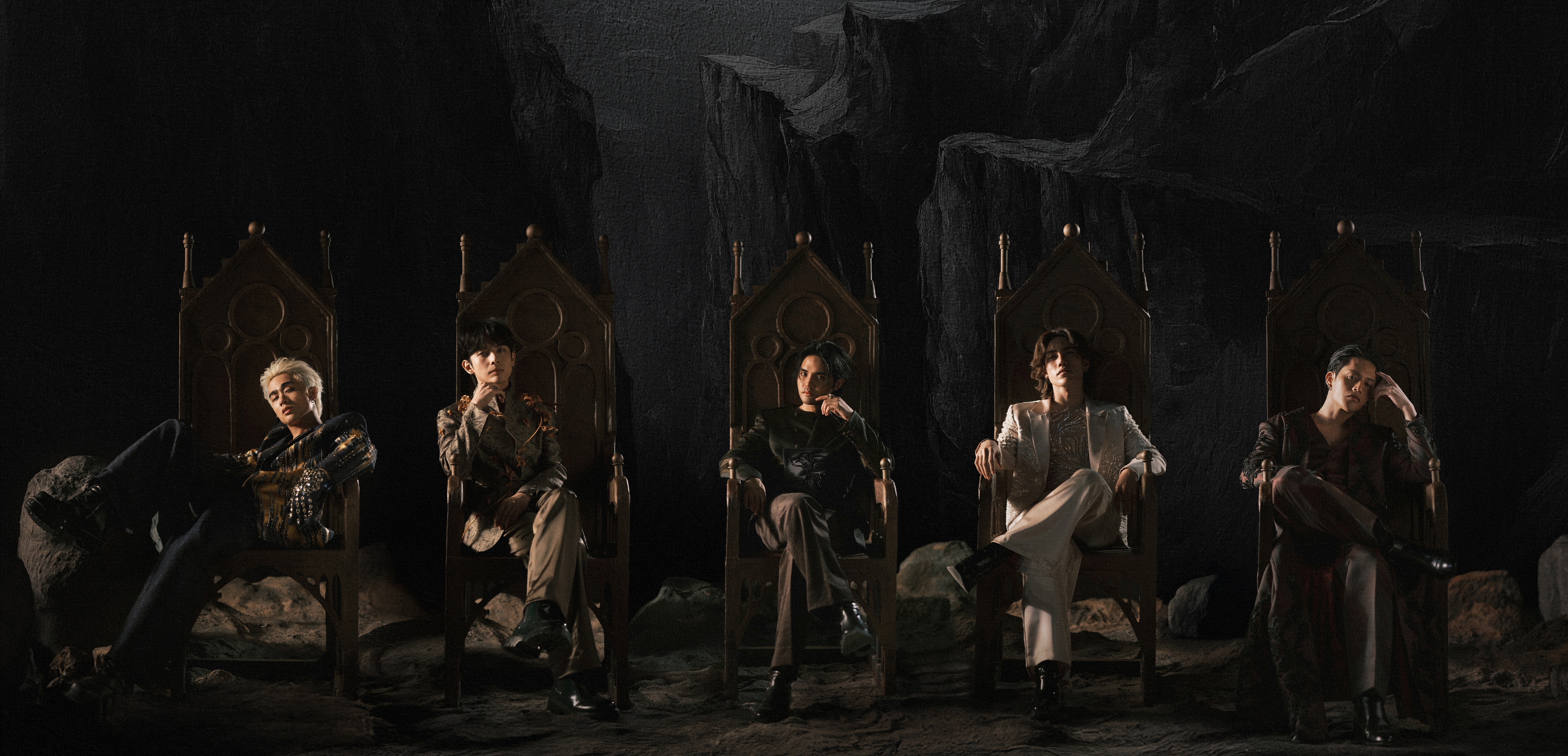 Five people sit on ornate chairs in a dramatic, shadowy setting. They wear stylish, dark-themed outfits, exuding an aura of mystery and elegance