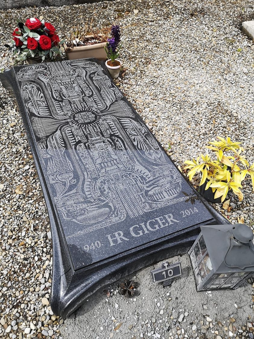 Artistic gravestone of H.R. Giger, engraved with elaborate biomechanical designs, surrounded by flowers and lamps