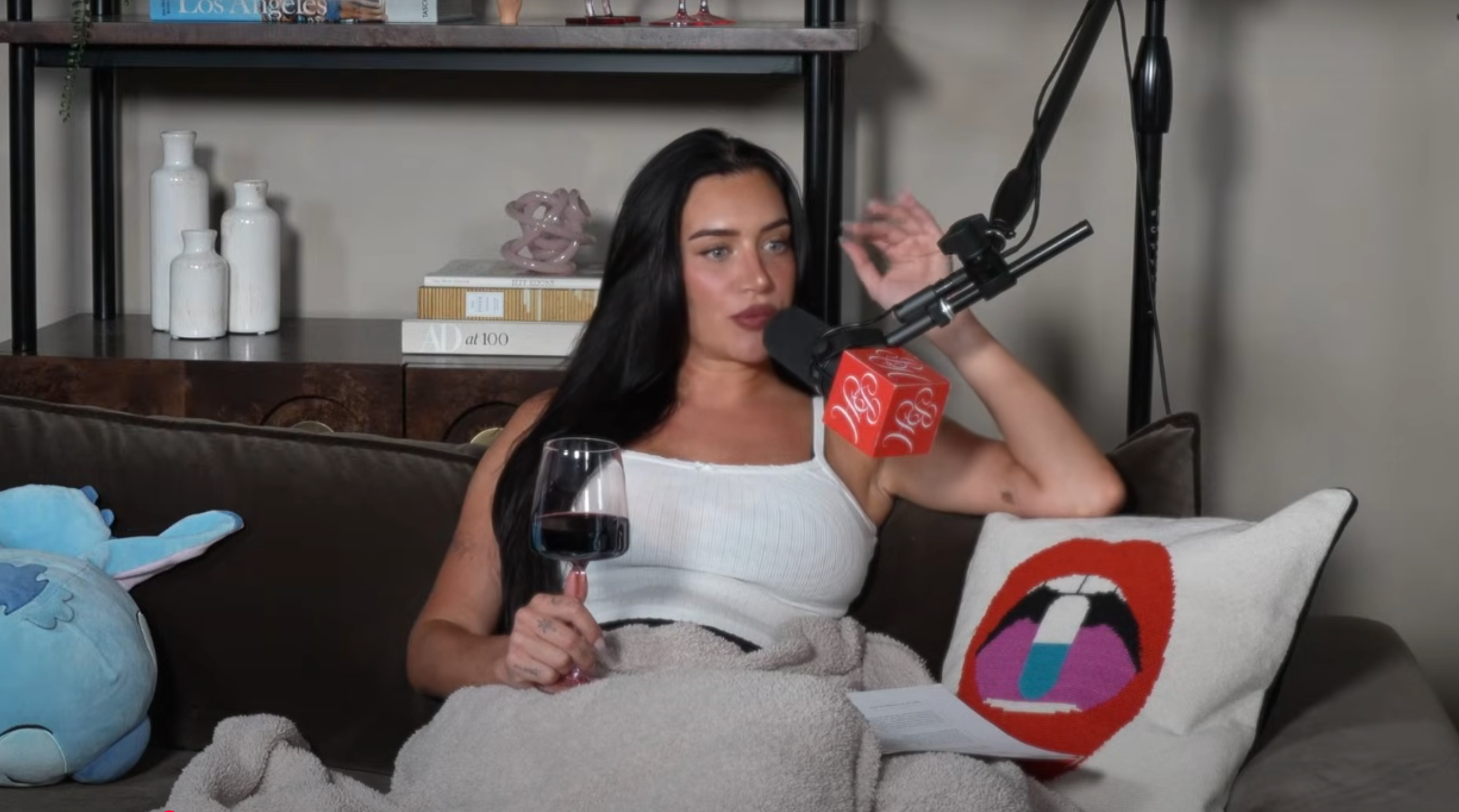 Person sitting on a couch holds a glass of wine, covered with a blanket, speaking into a microphone. A colorful pillow sits beside them