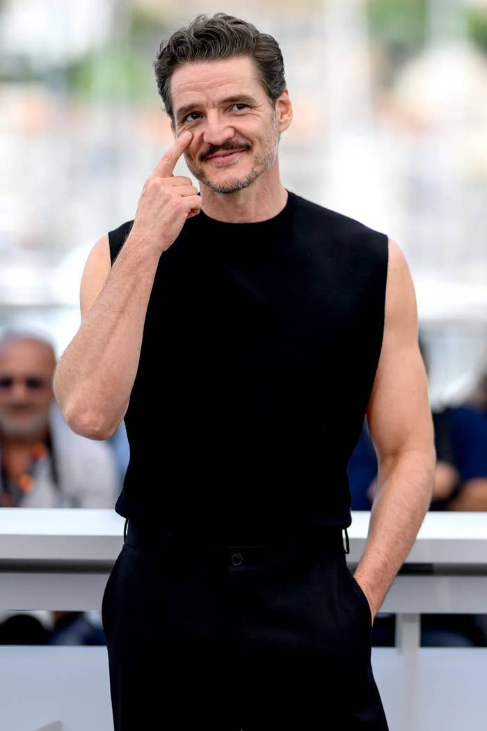 Persona con camisa sin mangas negra, sonriendo y señalando su mejilla, párase frente a una multitud, posiblemente en un evento público o festival