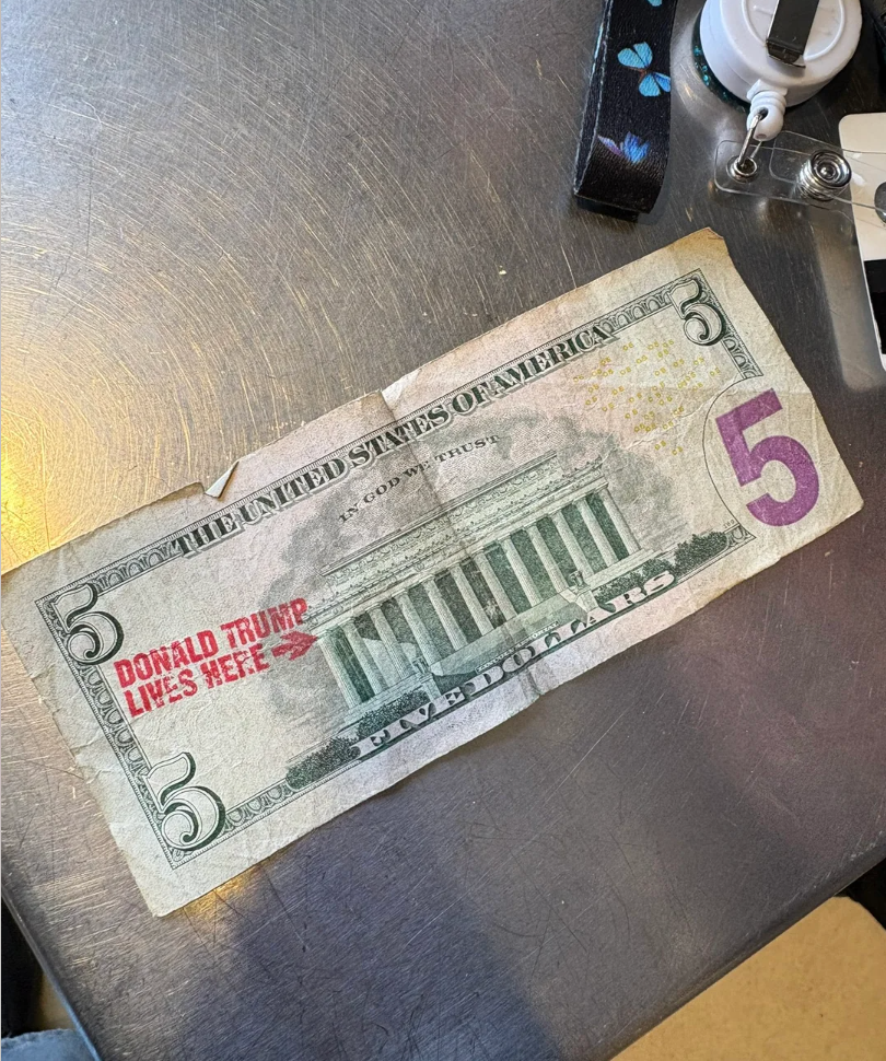 A five-dollar bill with a stamp reading "Donald Trump Lives Here" and an arrow pointing to the Lincoln Memorial