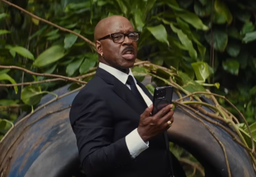 Hombre elegante con traje y gafas sosteniendo un teléfono frente a un fondo de vegetación, expresión seria
