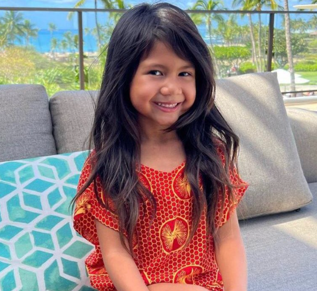 Una niña sonriente sentada en un sofá al aire libre, con un vestido de estampado llamativo