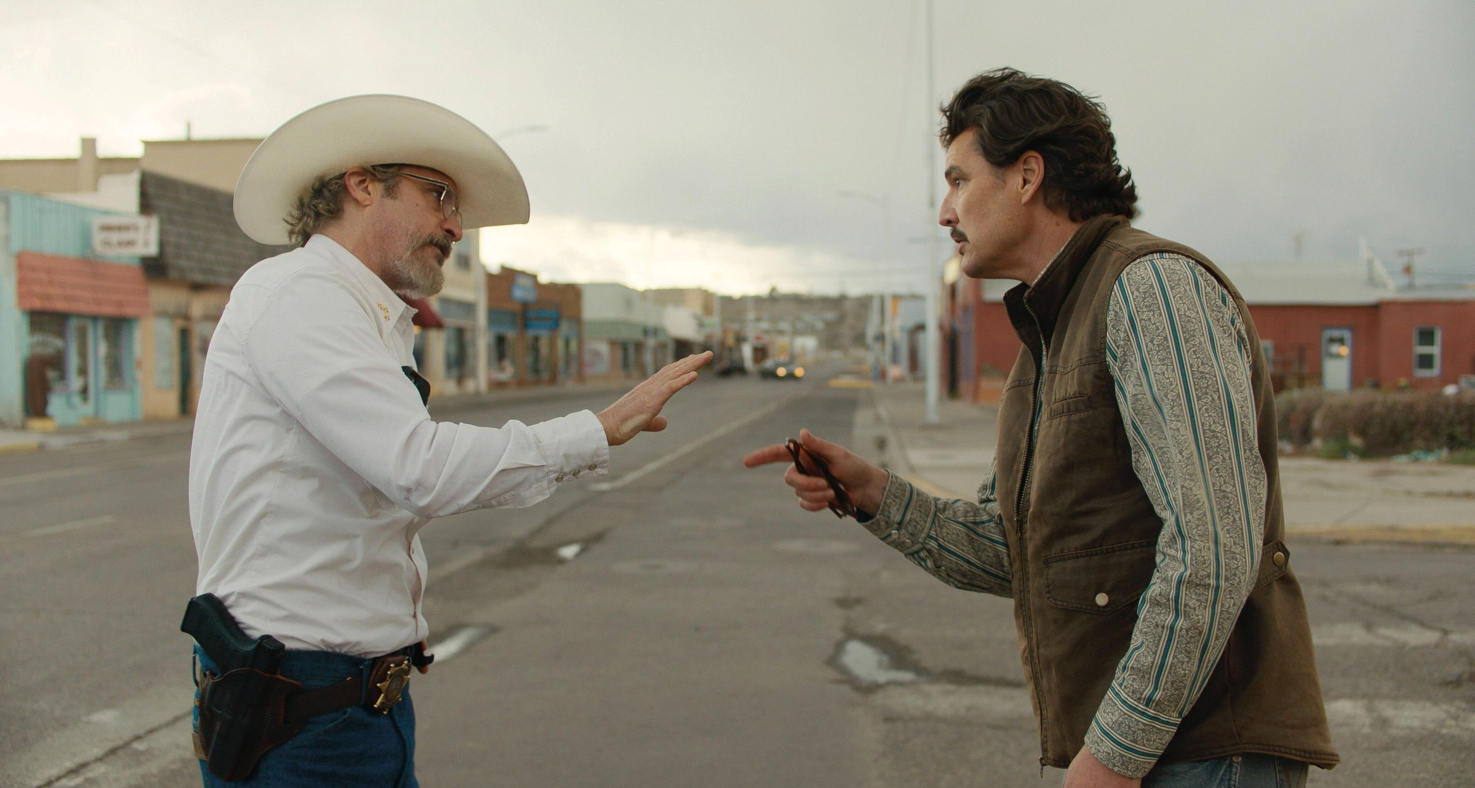 Two men stand in a street, engaging in a heated conversation. One wears a cowboy hat and jeans, the other a vest and patterned shirt