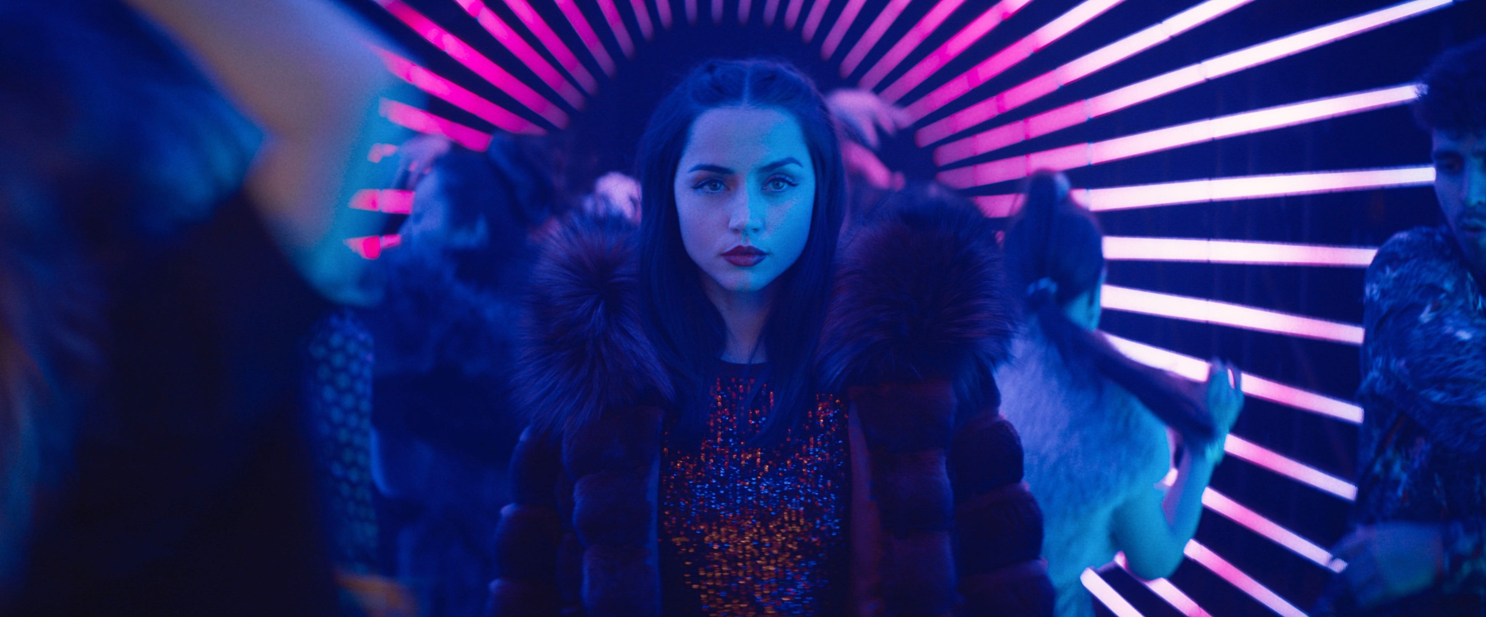 Person in a fur-collared coat stands confidently in a vibrant, neon-lit environment with people dancing around
