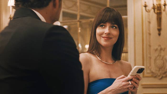 Woman in elegant, strapless dress smiling while looking at a man in a suit, holding a phone in an ornate room setting