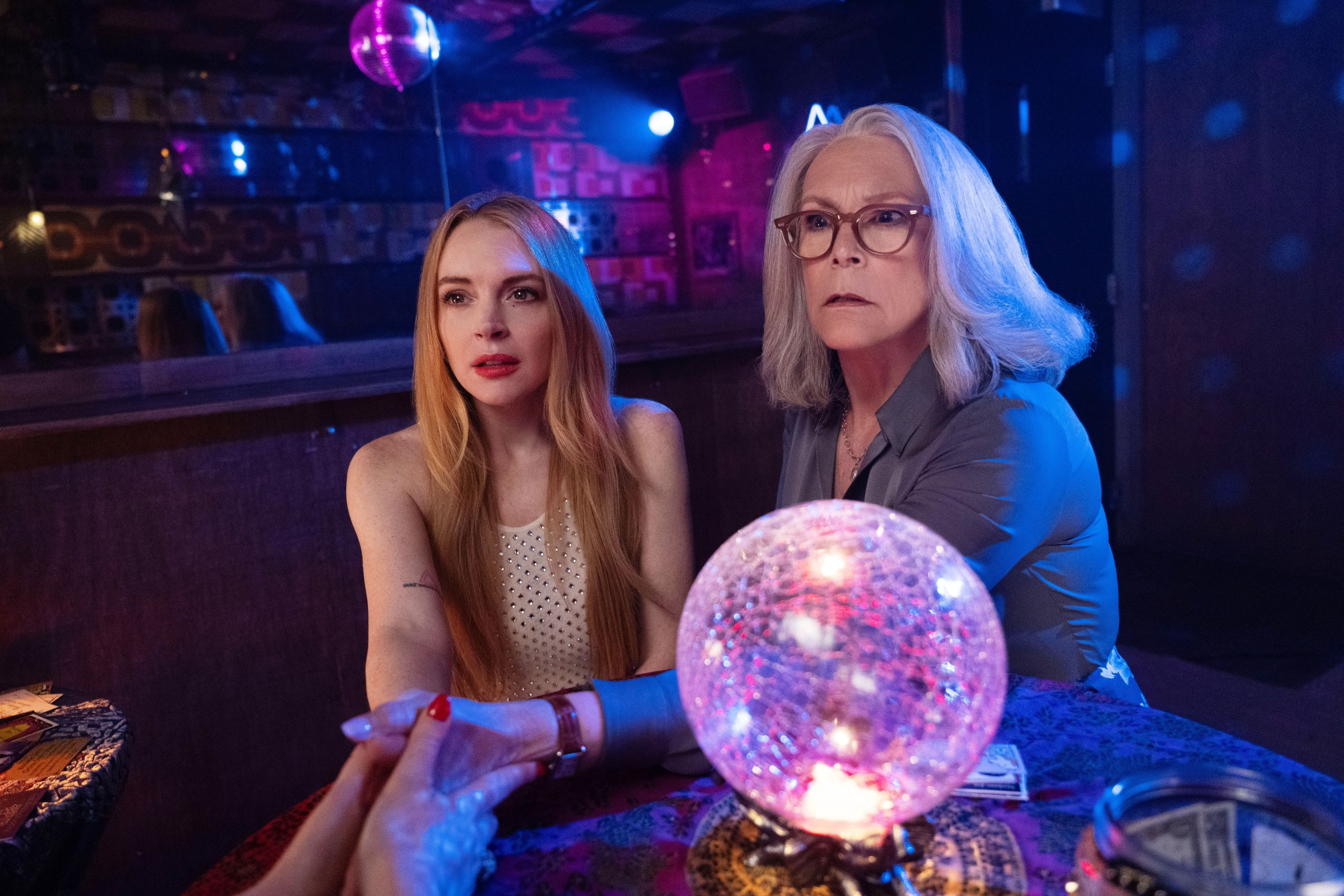 Two people sit at a table with a crystal ball in a dimly lit room, looking intrigued. One wears a sleeveless top, the other glasses and a long-sleeve shirt