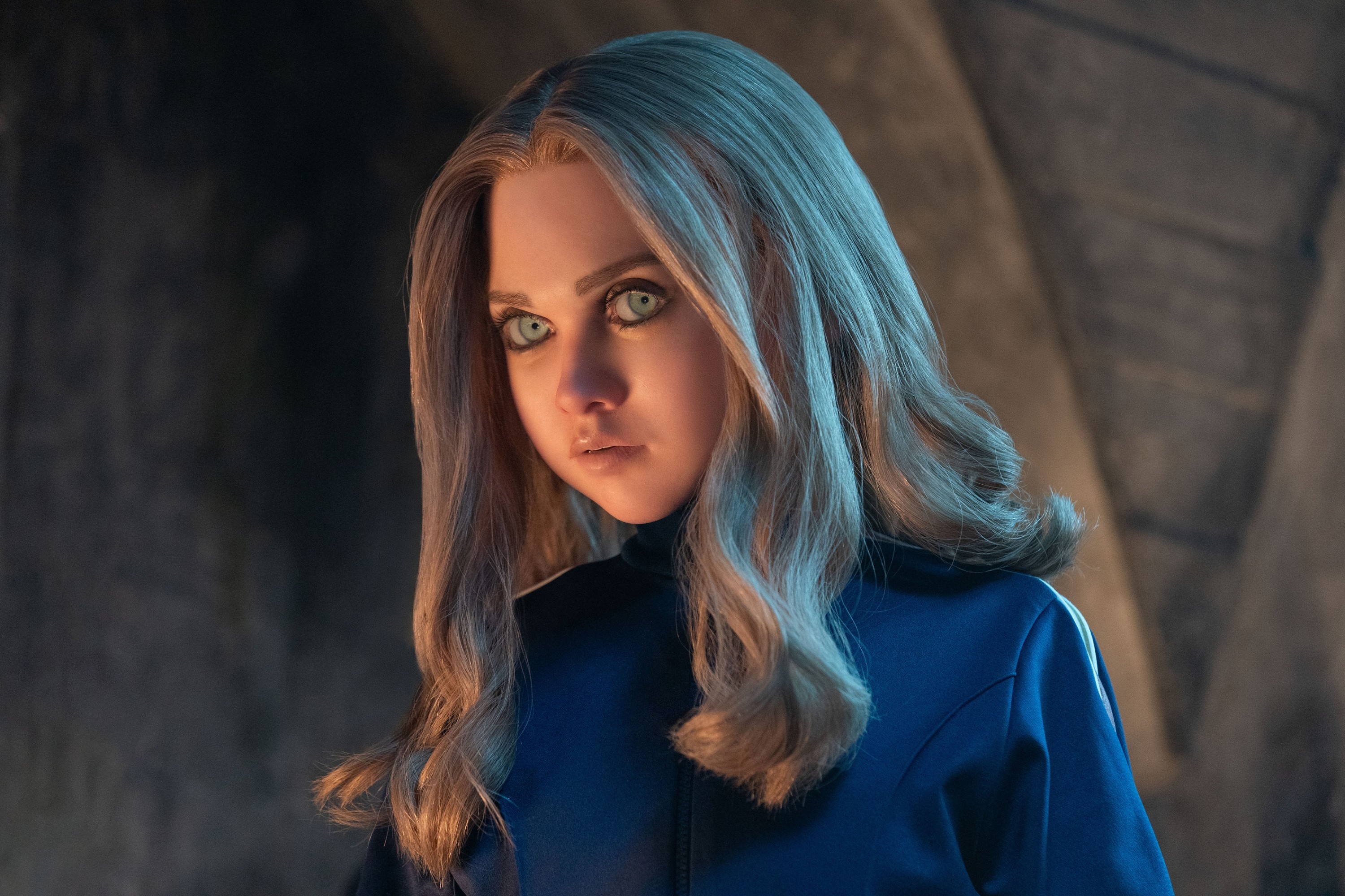 A lifelike humanoid character with long hair stares intently in a dimly lit setting, wearing a sleek outfit