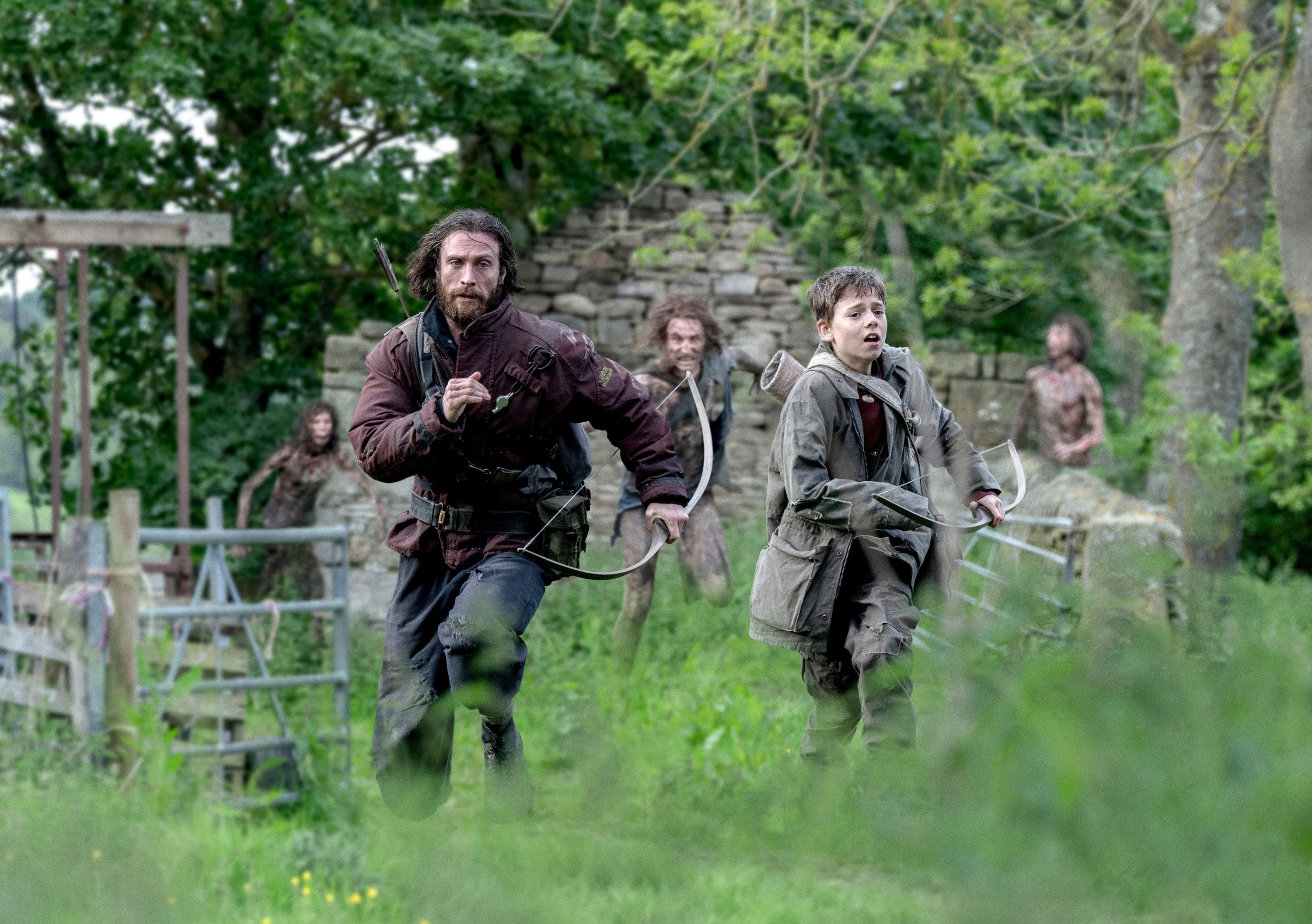 Characters in medieval-style clothing run through a grassy area, looking anxious, in a scene from a TV show or movie
