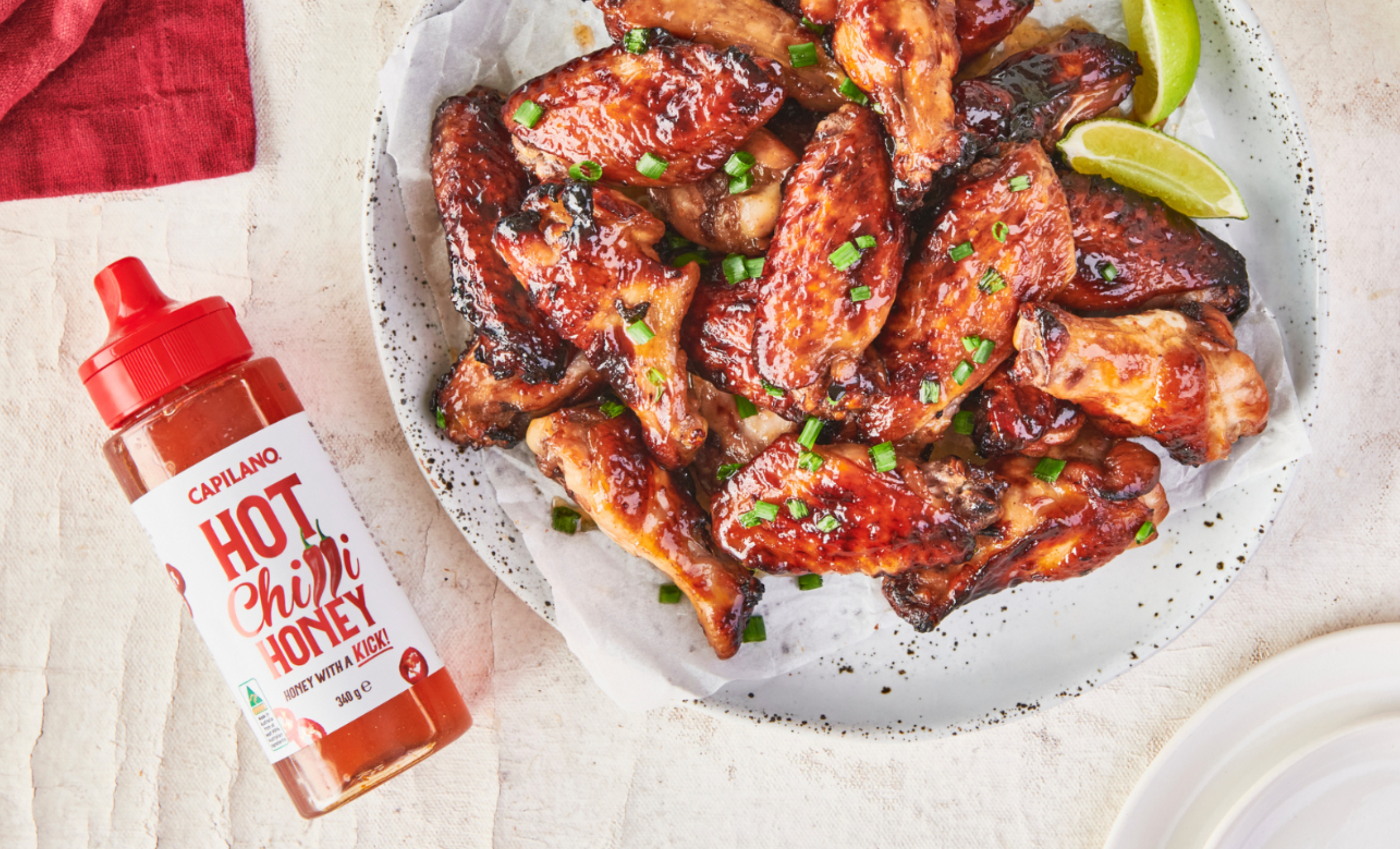 Plate of barbecued chicken wings garnished with herbs, next to a bottle of Capilano Hot Chilli Honey and lime wedges
