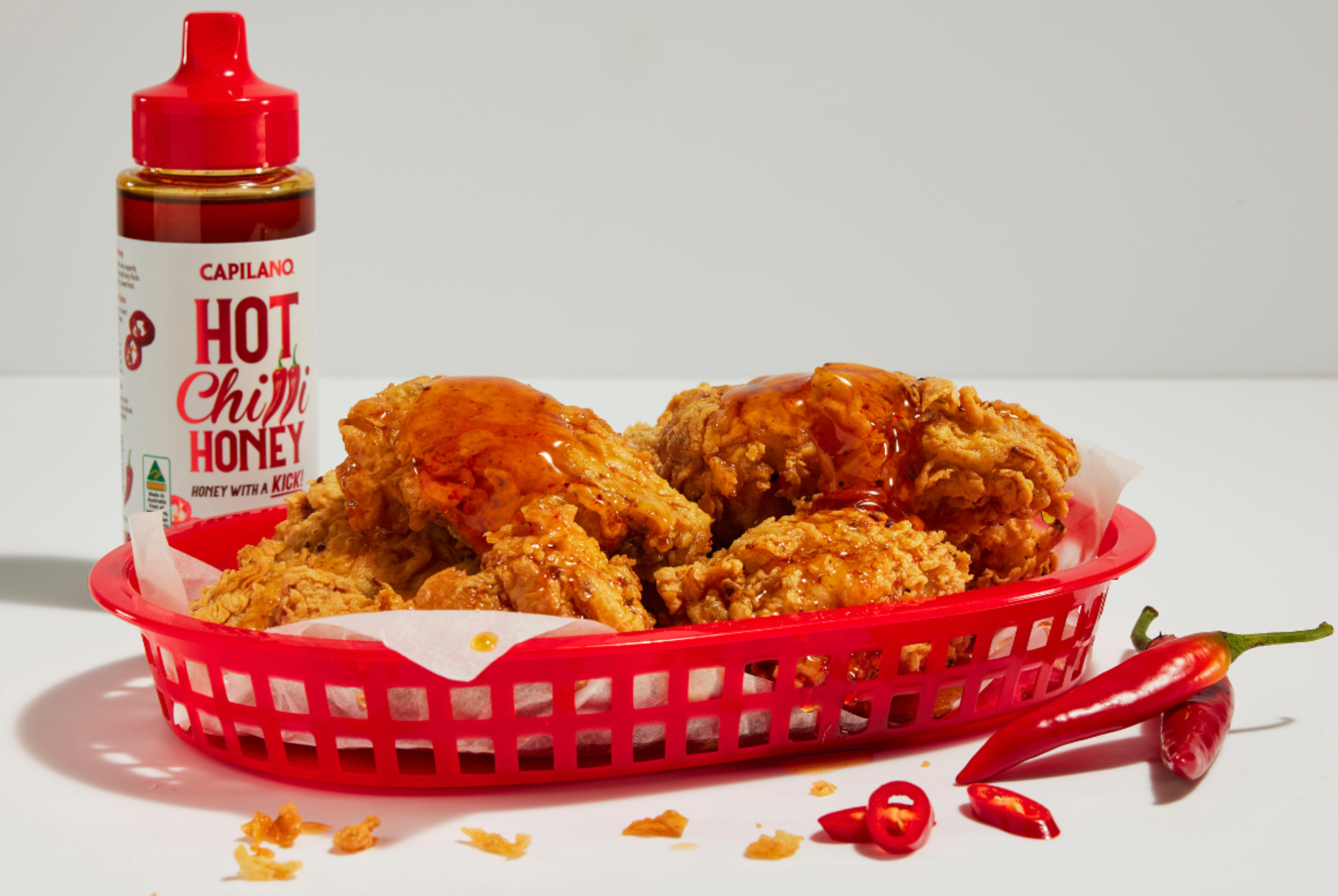 Fried chicken in a red basket is drizzled with Capilano Hot Chili Honey. A bottle and some chilies are also visible