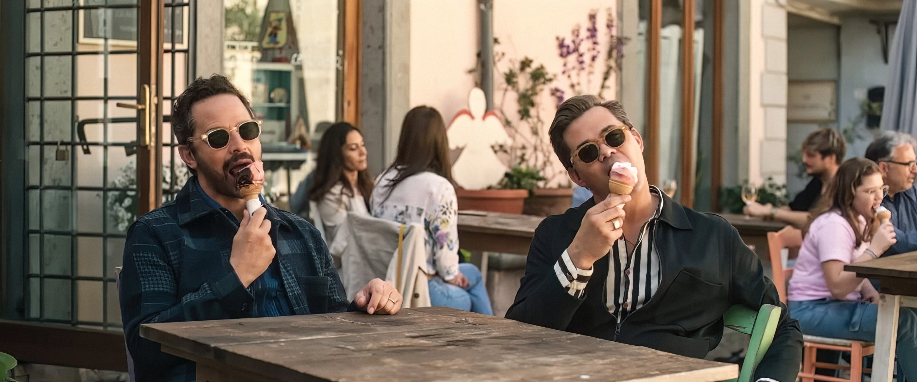 Two people in sunglasses casually sit outdoors, enjoying ice cream at a wooden table, with others in the background
