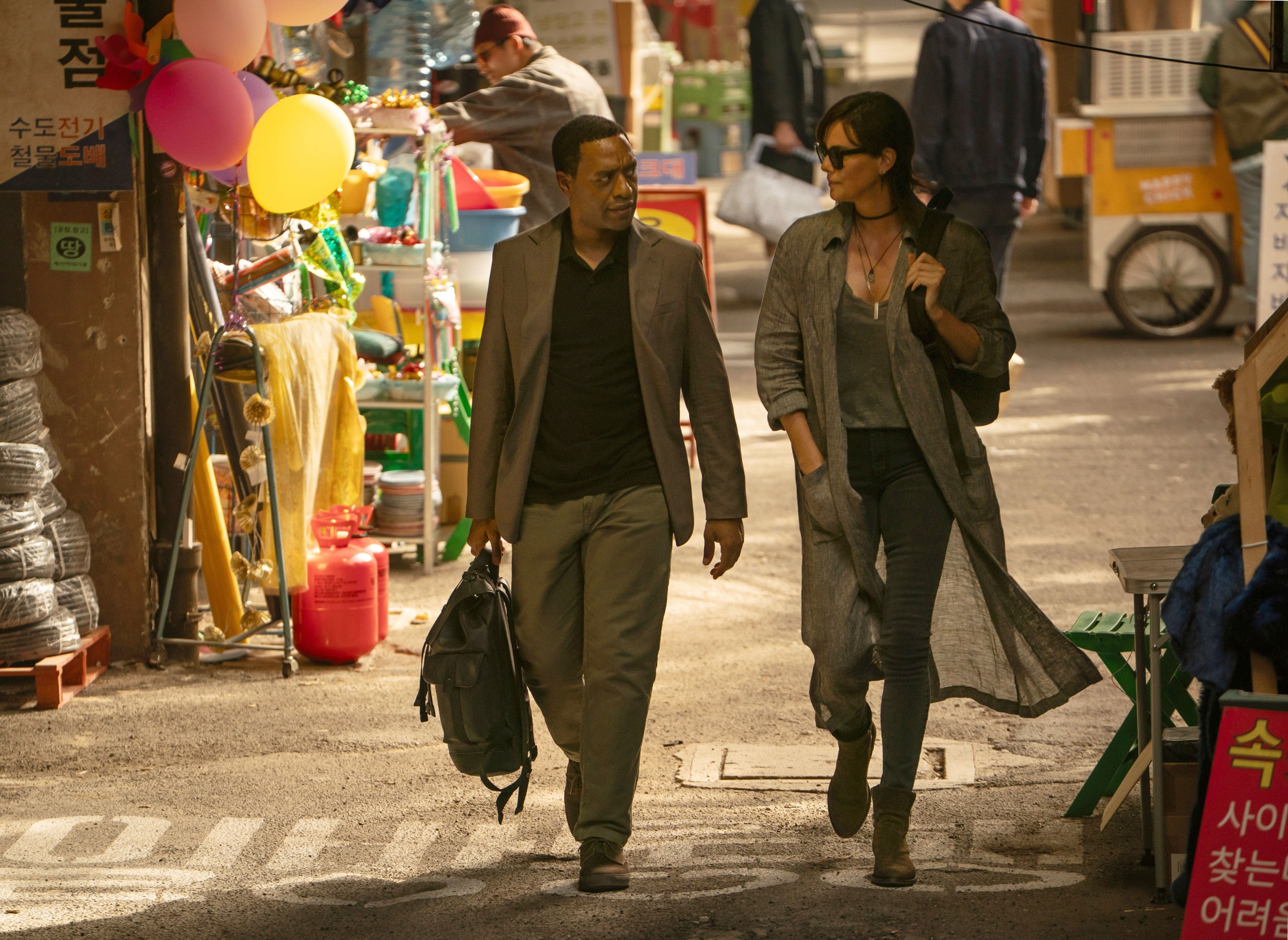 Two people walk through a bustling market, wearing casual yet stylish outfits. One carries a black bag, and they seem engaged in conversation