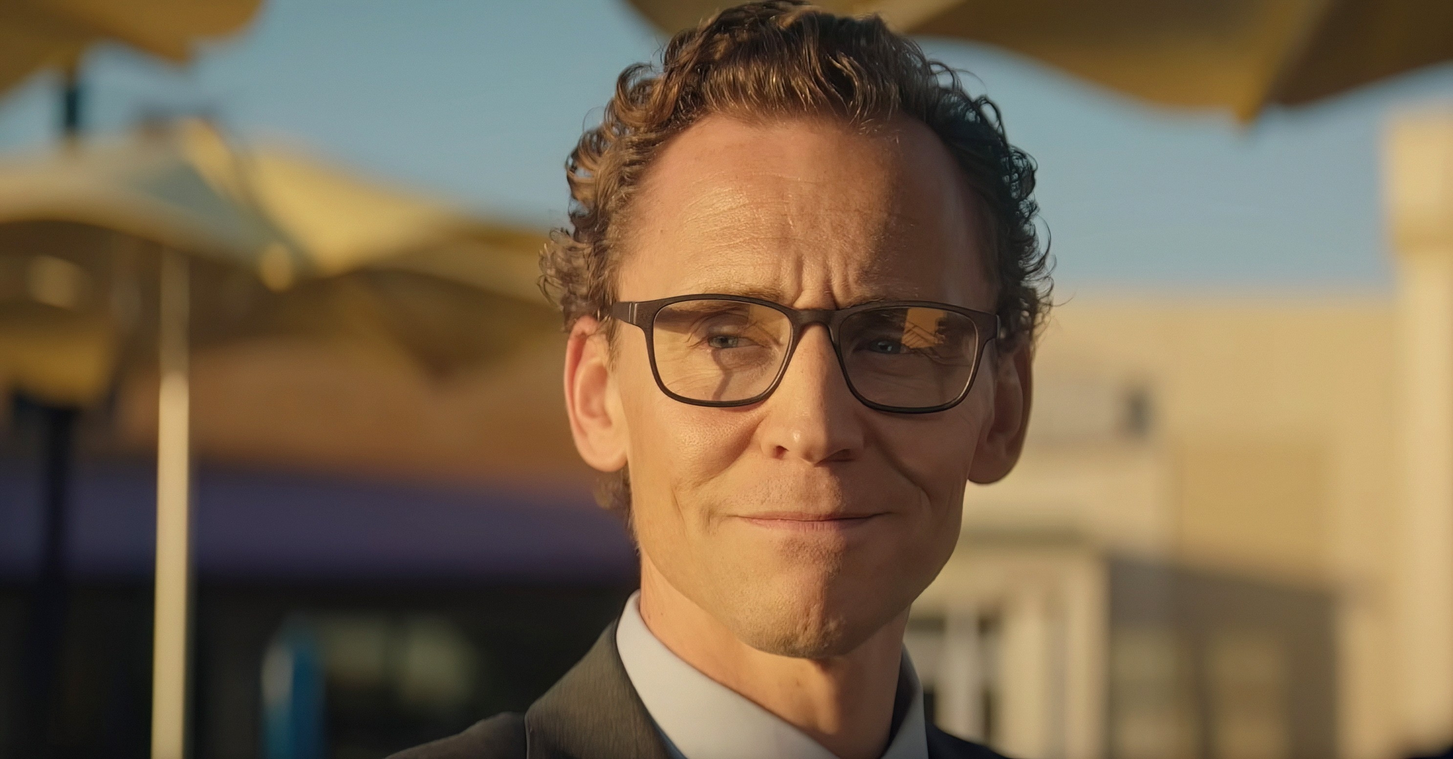 A person with curly hair and glasses smiles slightly, outdoors under umbrellas, wearing a suit