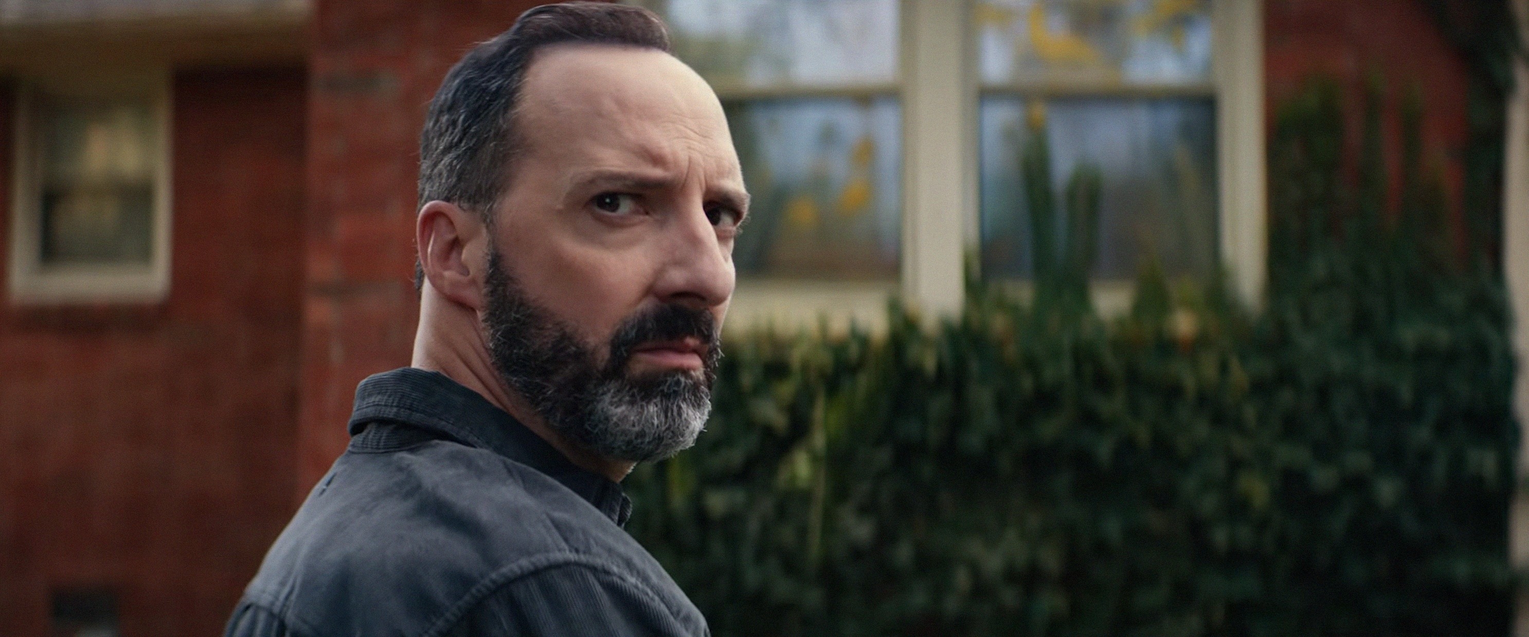 A man with a beard looks over his shoulder with a serious expression, standing in front of a brick house with greenery