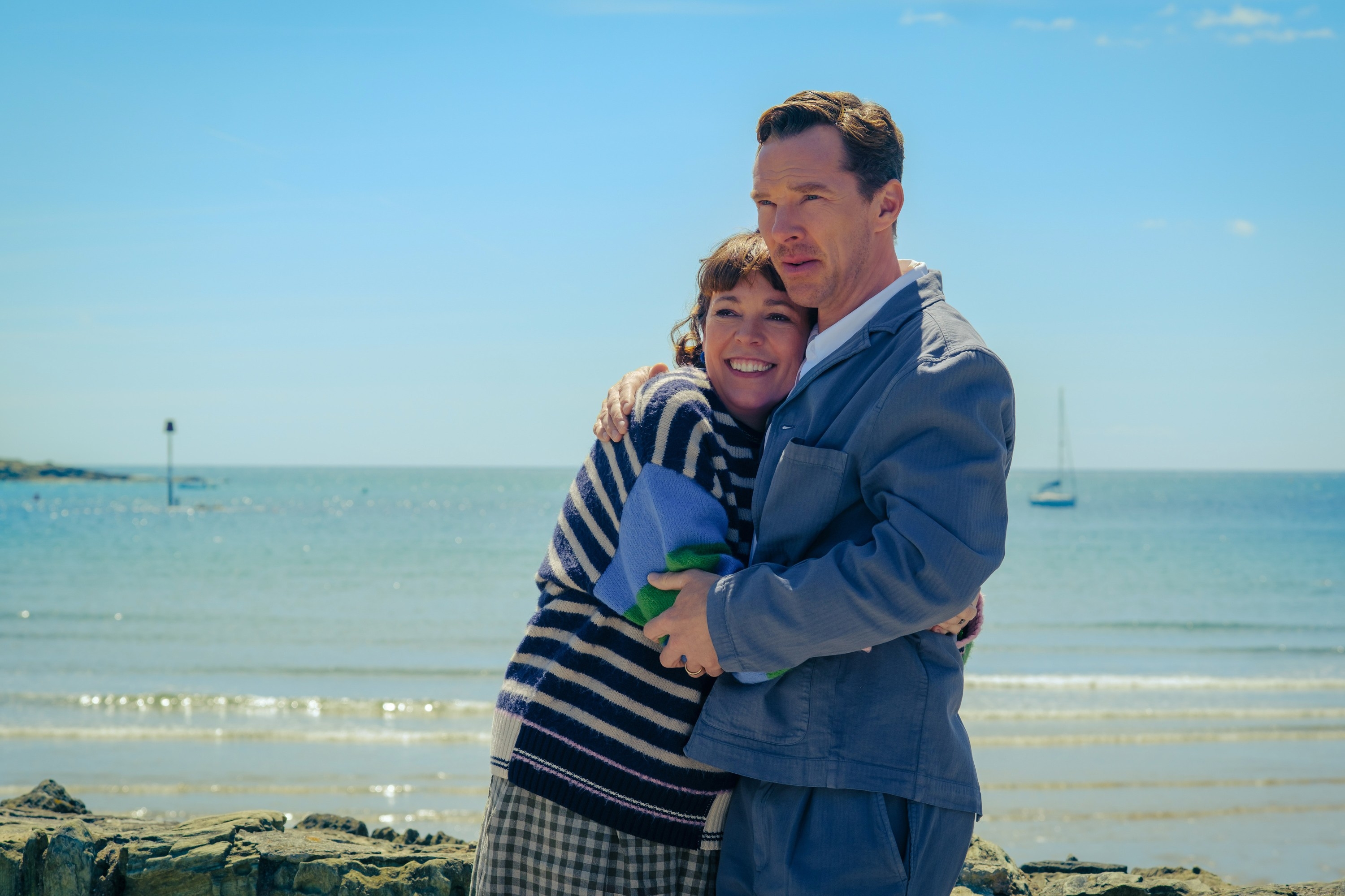 A man and woman embrace on a beach, both smiling. The man wears a suit, and the woman is in casual attire with a striped sweater