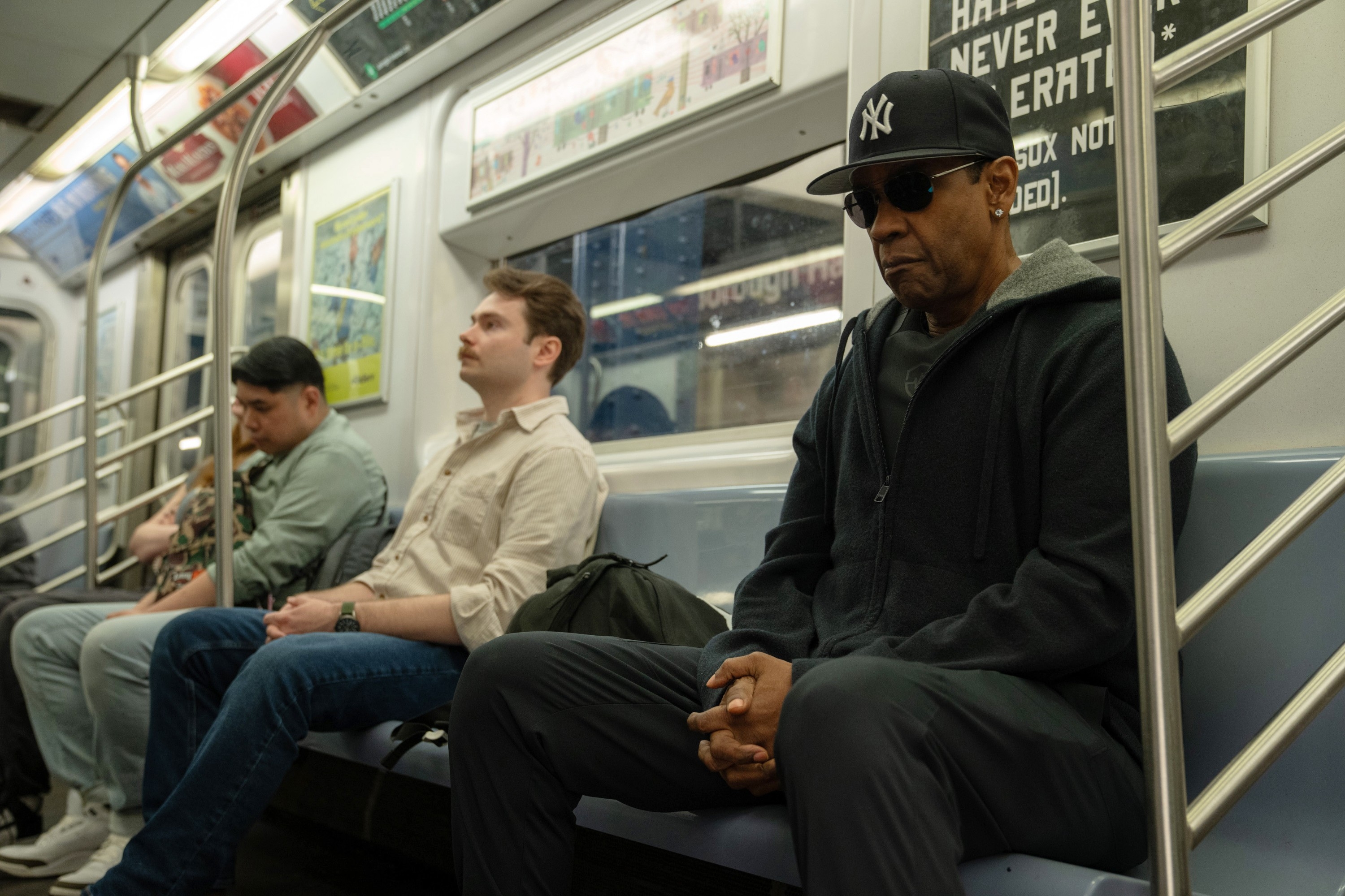 Three people sitting on a subway train bench. The person on the right wears a dark hoodie, sunglasses, and a cap