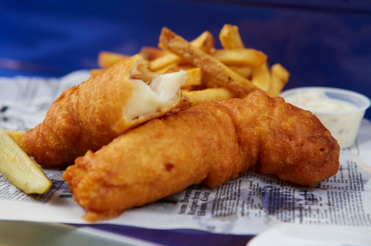 Fish and chips on newspaper with a lemon wedge and a small cup of tartar sauce in the background
