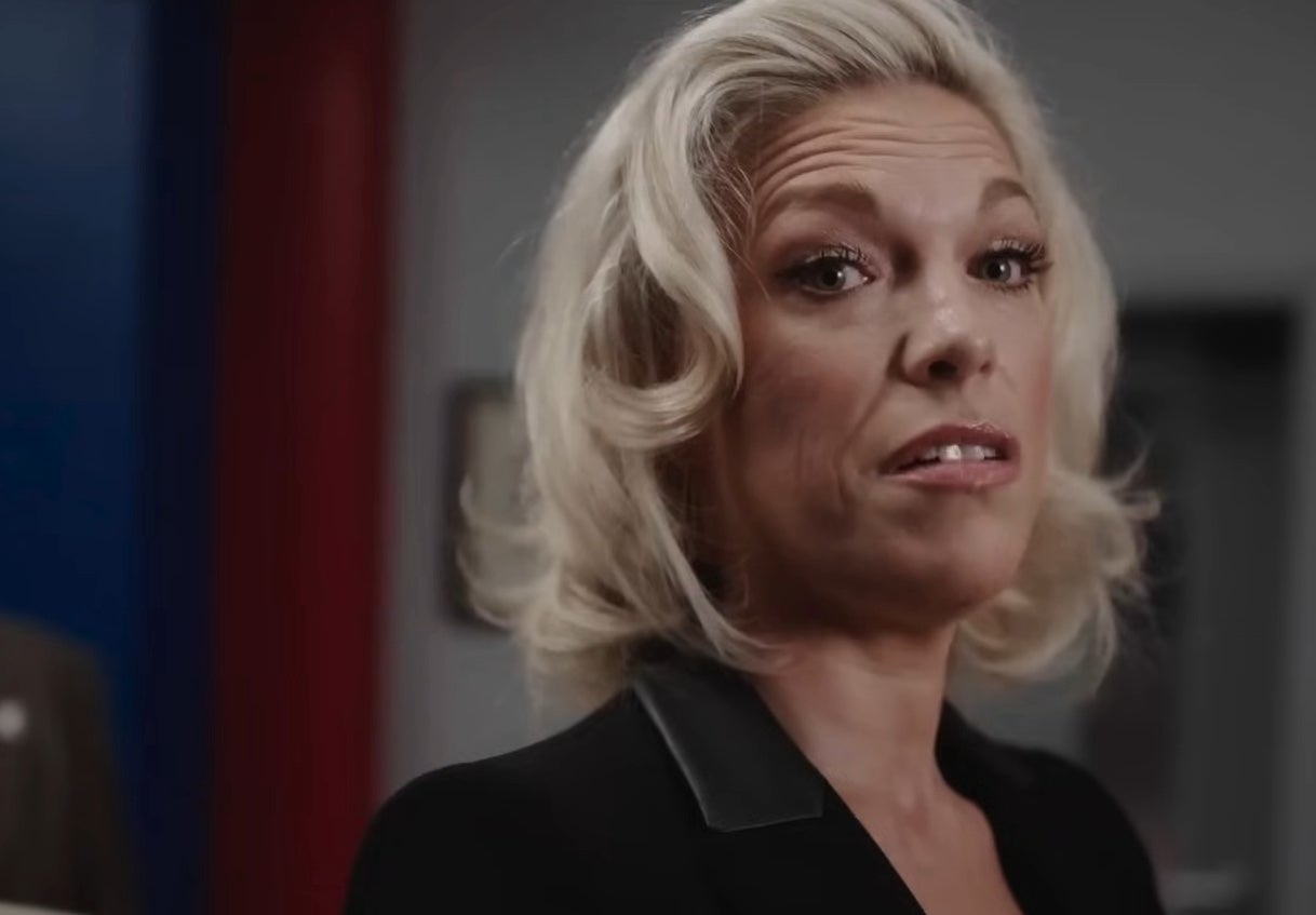 Person with short, wavy hair wearing a tailored blazer, glancing sideways with a slight expression during a formal event