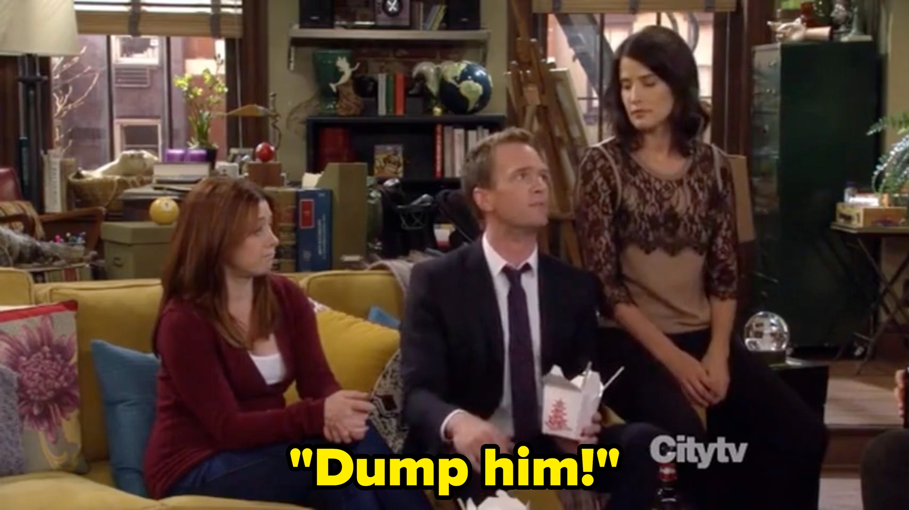 Three people sitting in a cozy living room, with open takeout boxes. The man wears a formal suit, and the two women wear casual tops