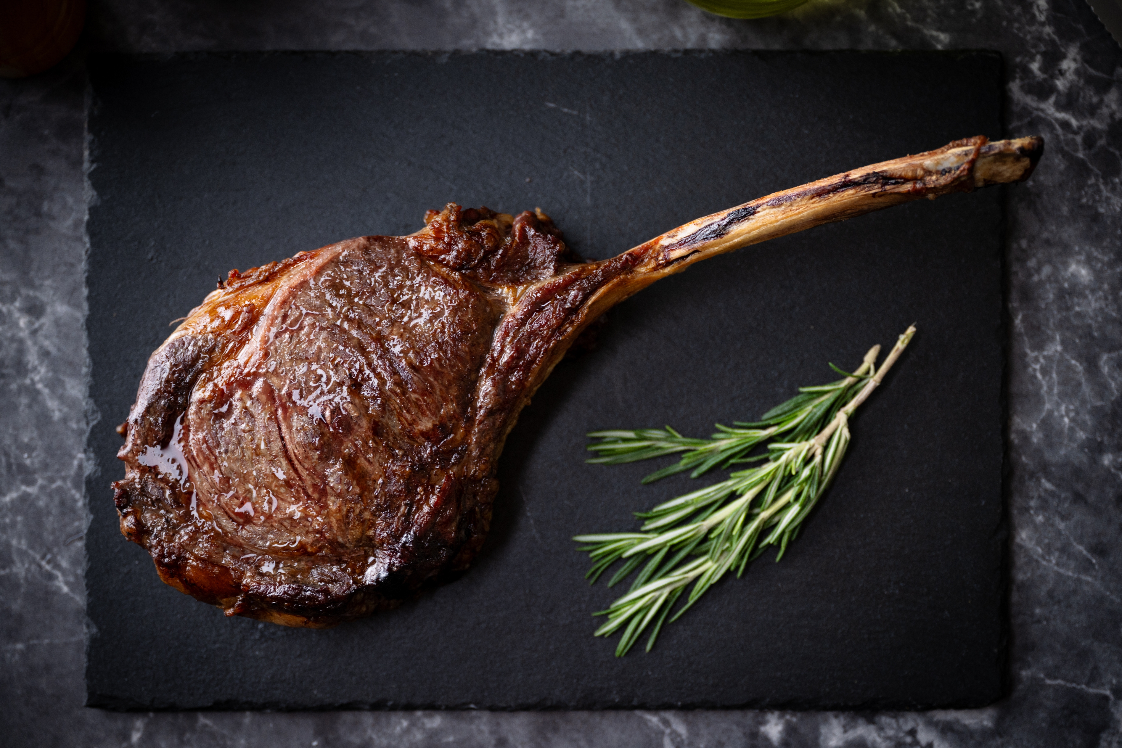 A cooked tomahawk steak on a black slate with a sprig of fresh rosemary beside it