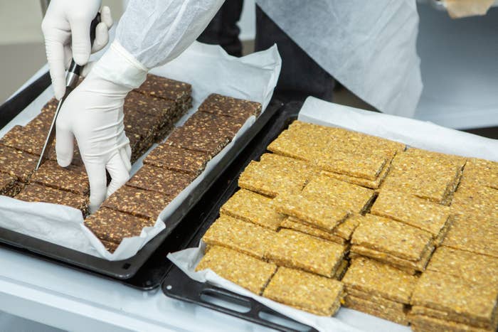 Gloved hands cut and arrange brown and yellow-brown bars on trays, possibly preparing or packaging food bars in a production setting