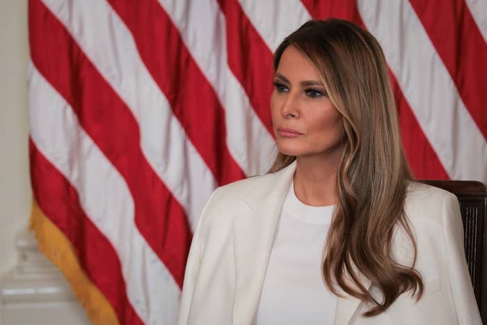 Melania Trump sits in front of a large American flag, looking thoughtfully to the side