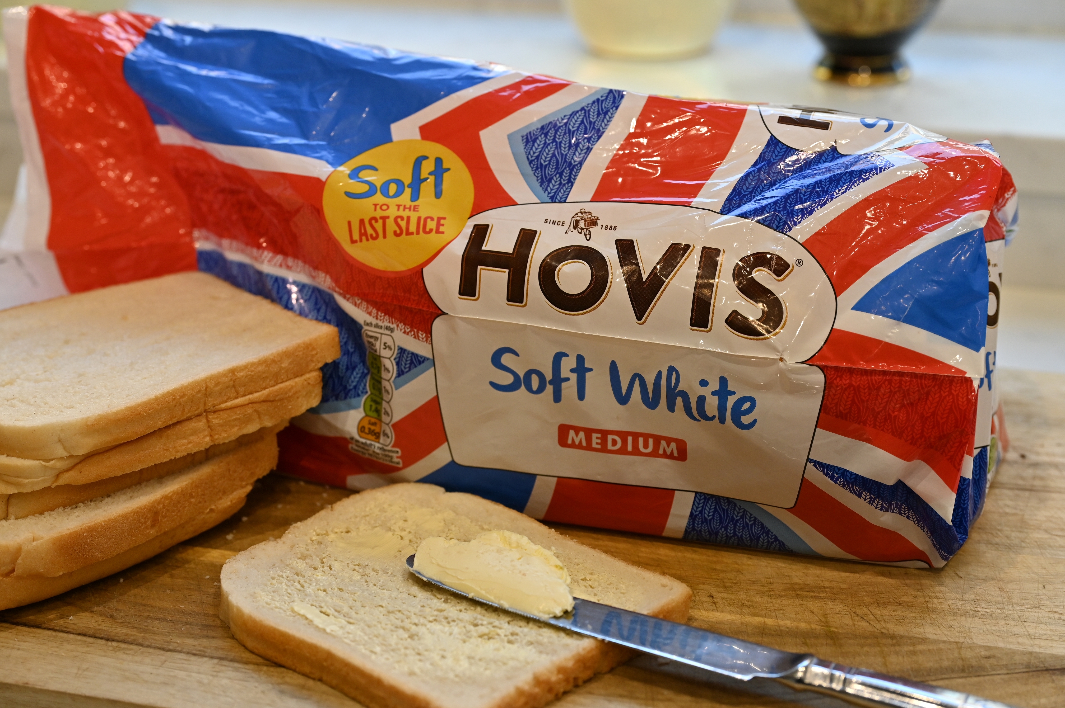 Pack of Hovis Soft White Medium sliced bread on a wooden board with a knife spreading butter on a slice. Bread packaging features a UK flag design