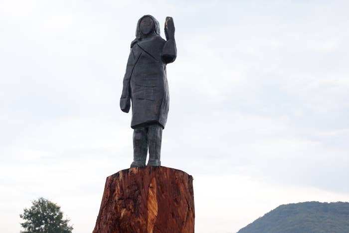 Bronze statue of a standing figure with one arm raised, carved atop a tree stump, against a backdrop of hills and cloudy sky
