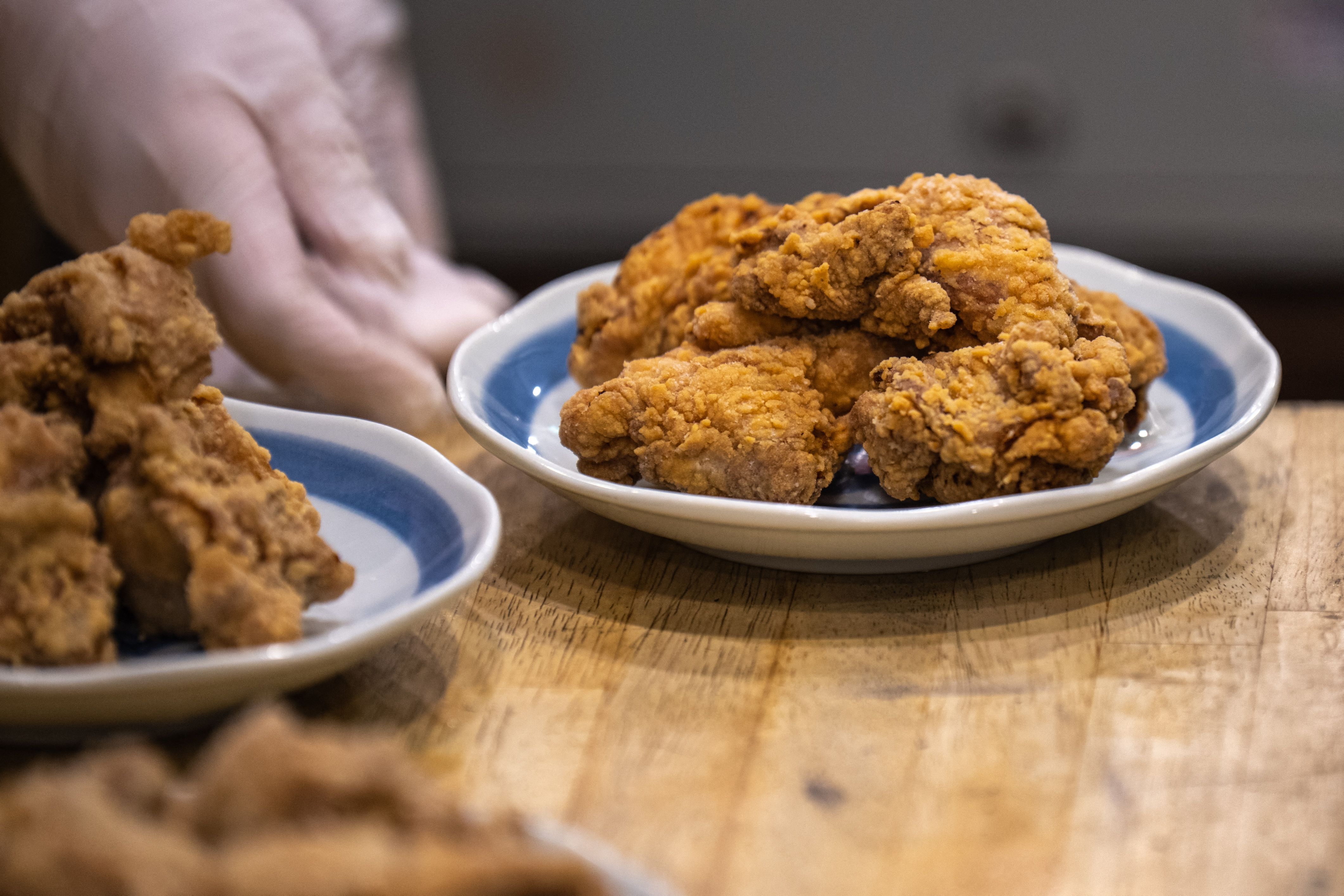 A plate of crispy fried chicken pieces is being served on a wooden table, with a gloved hand placing a dish nearby