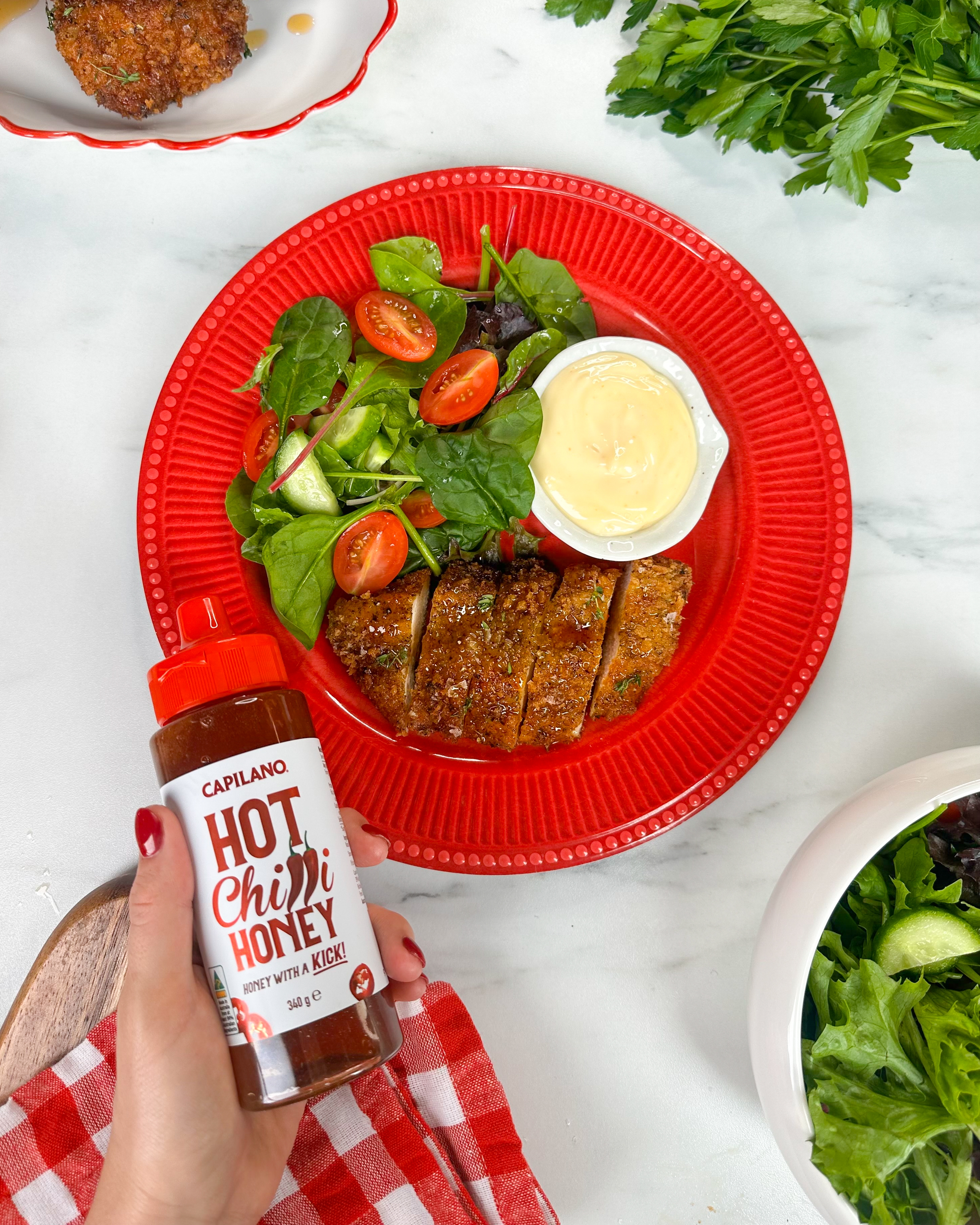 Plate of salad with sliced chicken and dressing beside a hand holding a bottle of Capilano Hot Chili Honey