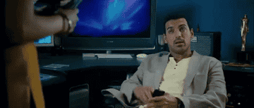 Man in a light suit and yellow shirt sits in an office chair, looking surprised and holding a phone. A woman stands nearby, out of focus