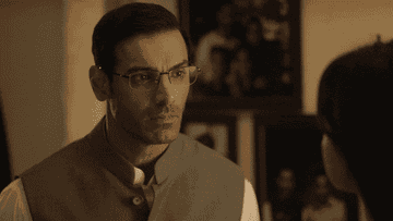 Man in glasses and traditional vest looks seriously at someone, standing in a warmly lit room with framed photos on the wall