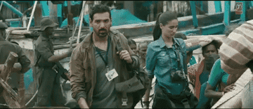 People walking through a crowded, rustic area with boats; wearing casual, expedition-style clothes with cameras and gear